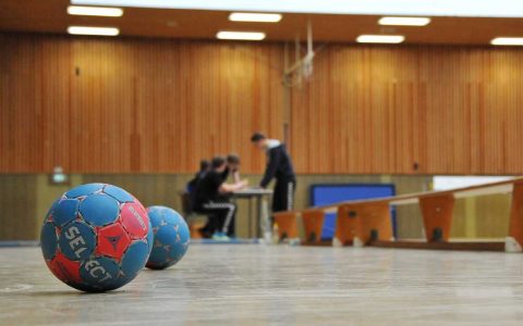 Handball im Vordergrund, hinten Vereinsmitglieder an einem Tisch stehend in einer Sporthalle. Energiekostenzuschüsse helfen, dass das Licht nicht aus geht.